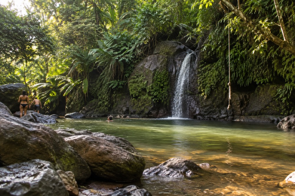 Afternoon sun over a broad river pool with a thin waterfall spilling off a mossy rock wall