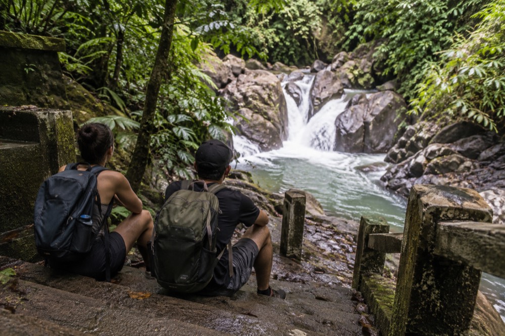 Hikers with backpacks resting on a mossy stone step beside a rainforest stream