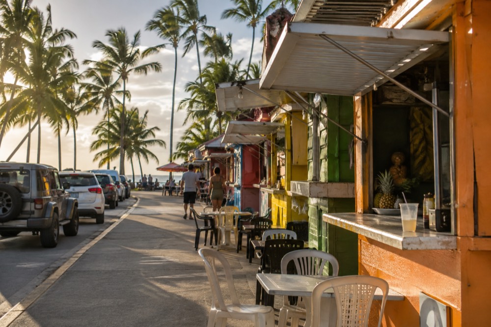 A row of colorful food kiosks beside a palm-lined coastal road