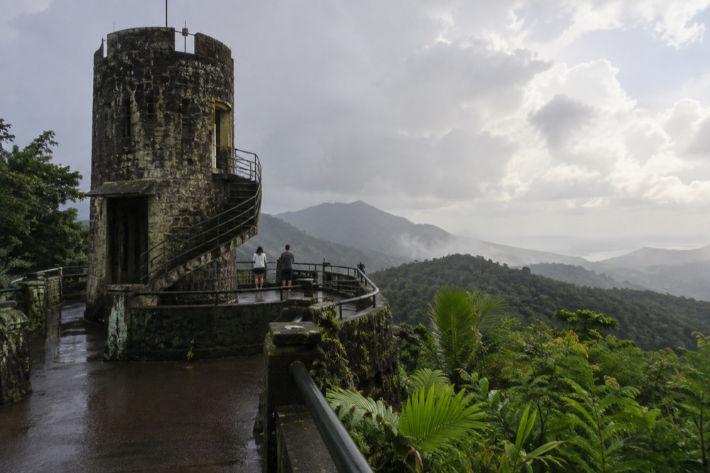 Stone observation tower above layered green mountain ridges under a hazy sky.