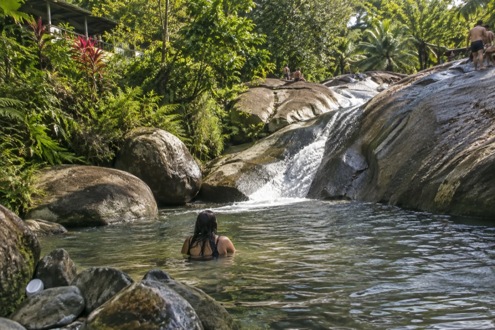 A shallow river pool at the base of a small waterfall surrounded by large smooth rocks and green trees