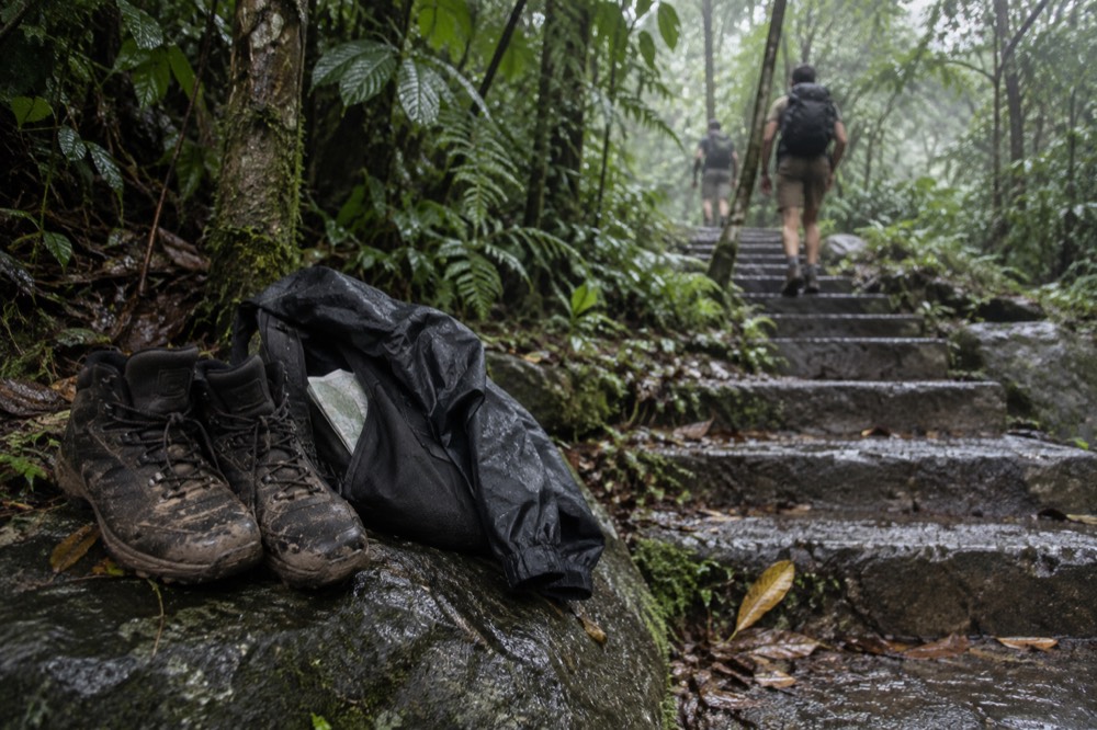 Hiking boots and a rain jacket resting on a wet rock beside a forest trail