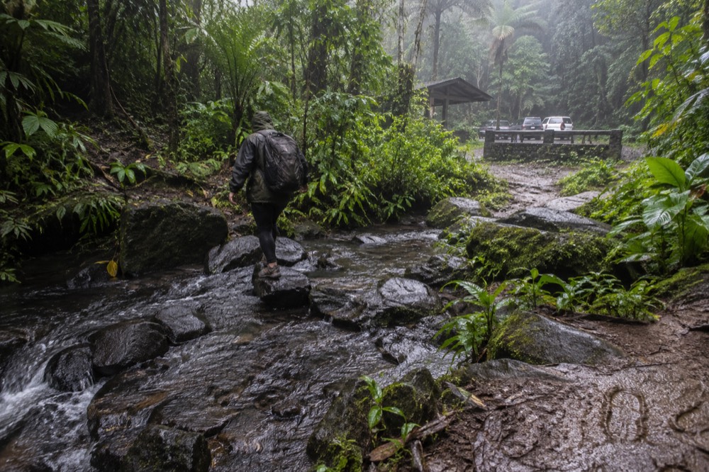 A hiker in a rain jacket crossing a shallow mountain stream on wet rocks
