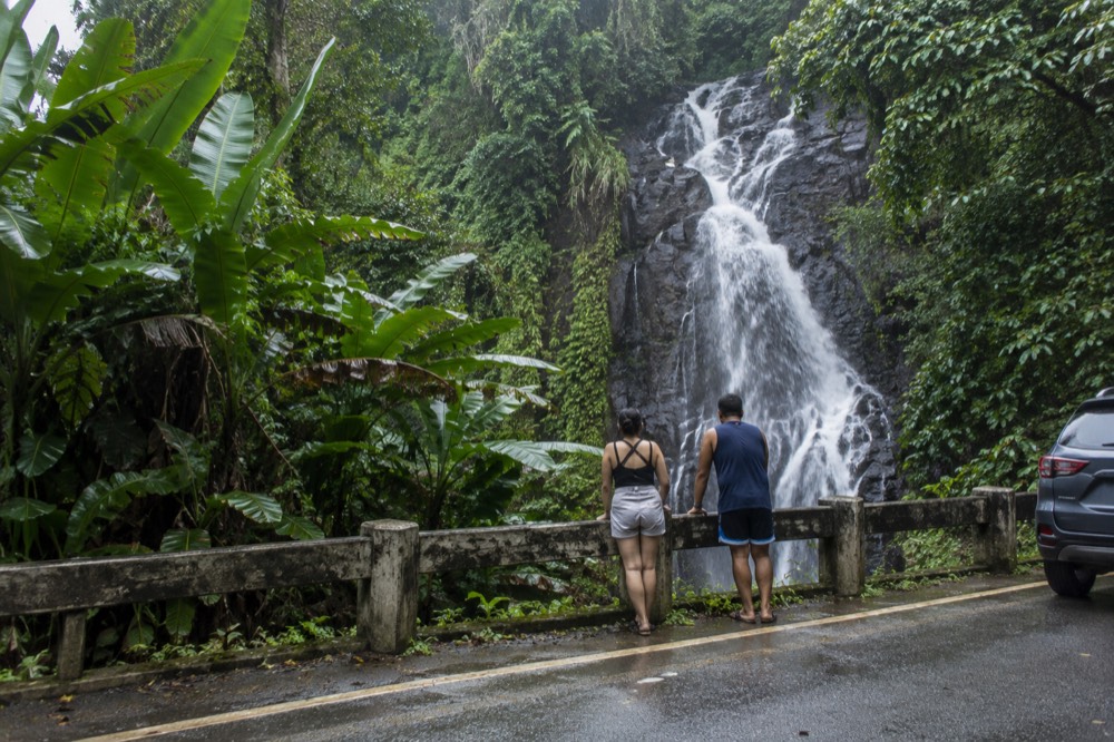 A waterfall seen from a forest road pull-off with dense tropical greenery