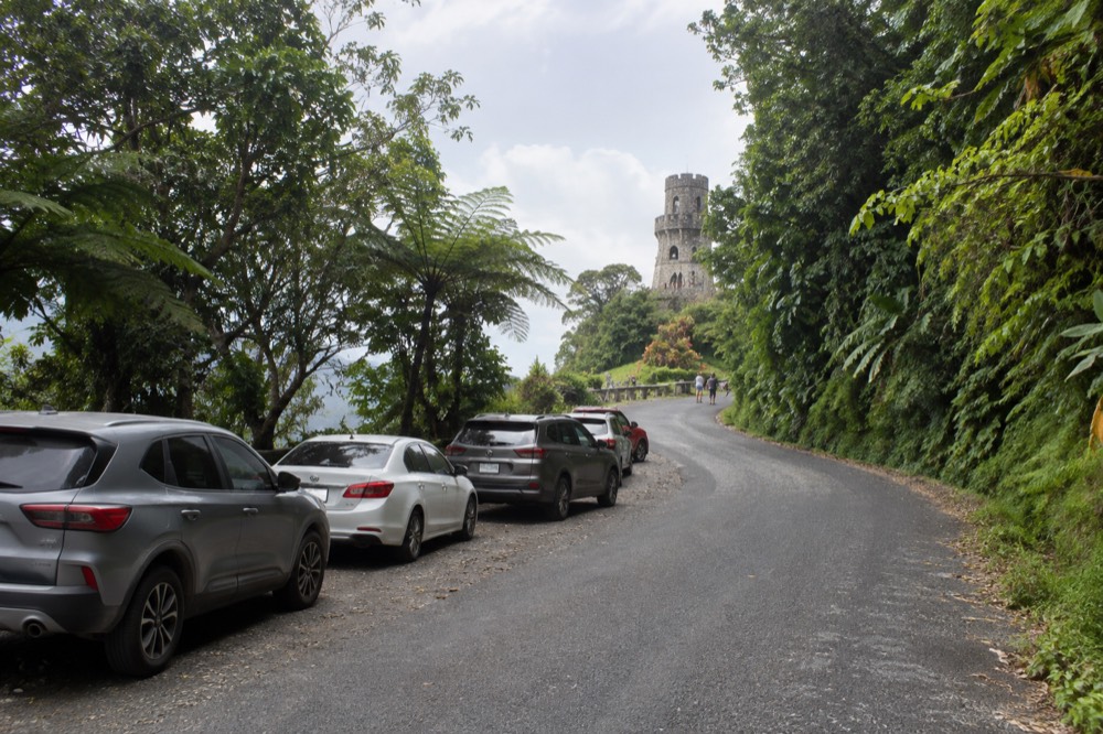 A forest road winding uphill past thick green vegetation with parked cars along a designated area