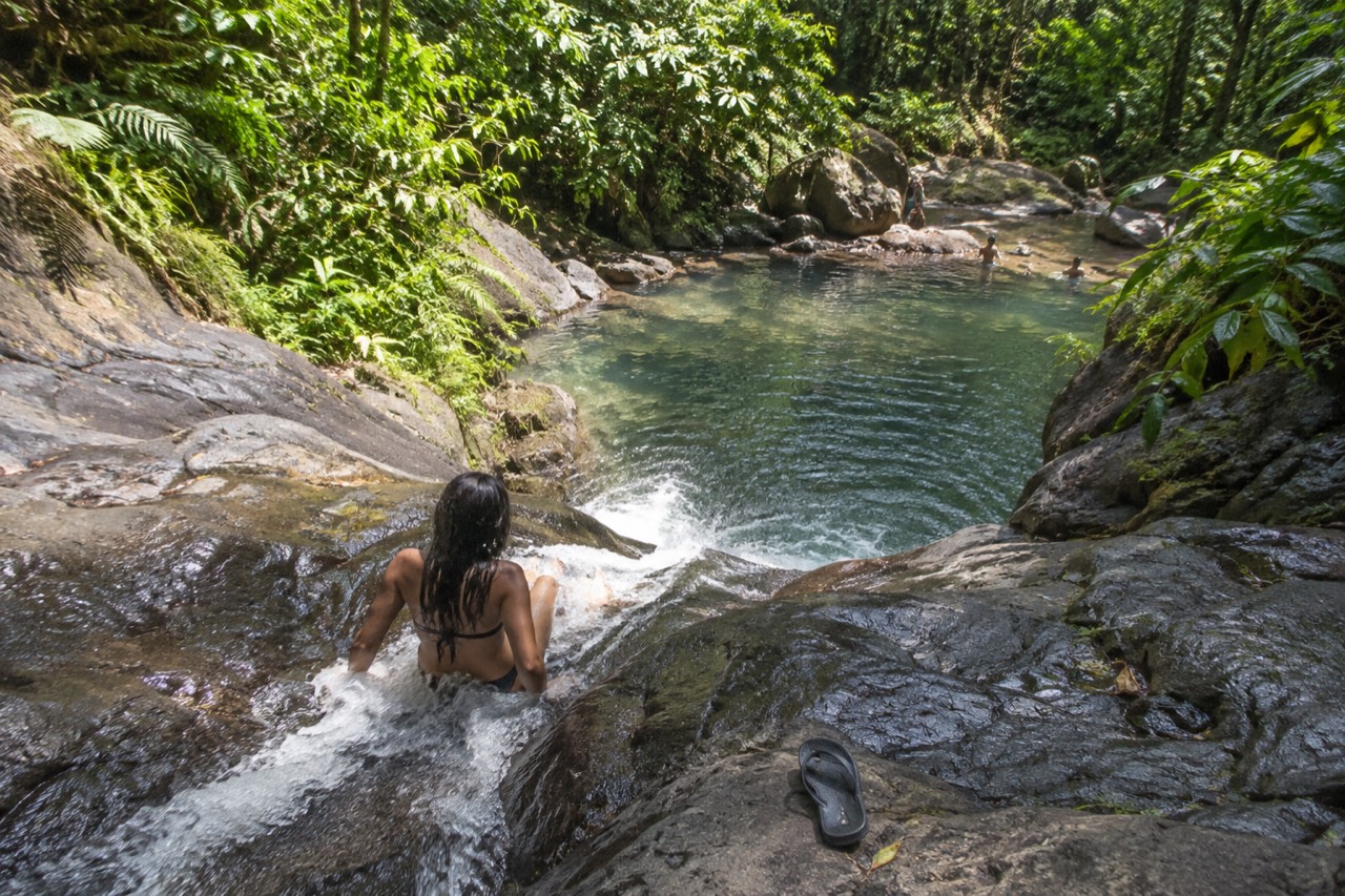 A smooth rock waterslide channel dropping into a clear jungle pool surrounded by green forest