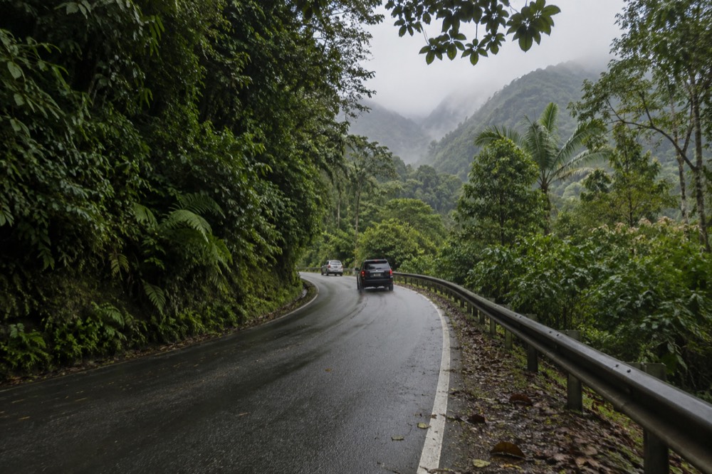 A winding mountain road climbing through dense tropical vegetation toward misty rainforest peaks