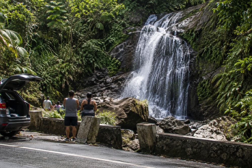 A tall waterfall spilling over a dark rock face beside a rainforest road with green foliage all around