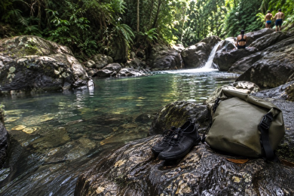Water shoes and a dry bag resting on a rock beside a shallow rainforest river