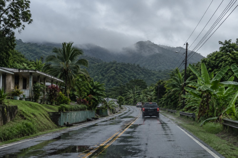 A two-lane mountain road climbing through dense rainforest with wet pavement and low clouds overhead