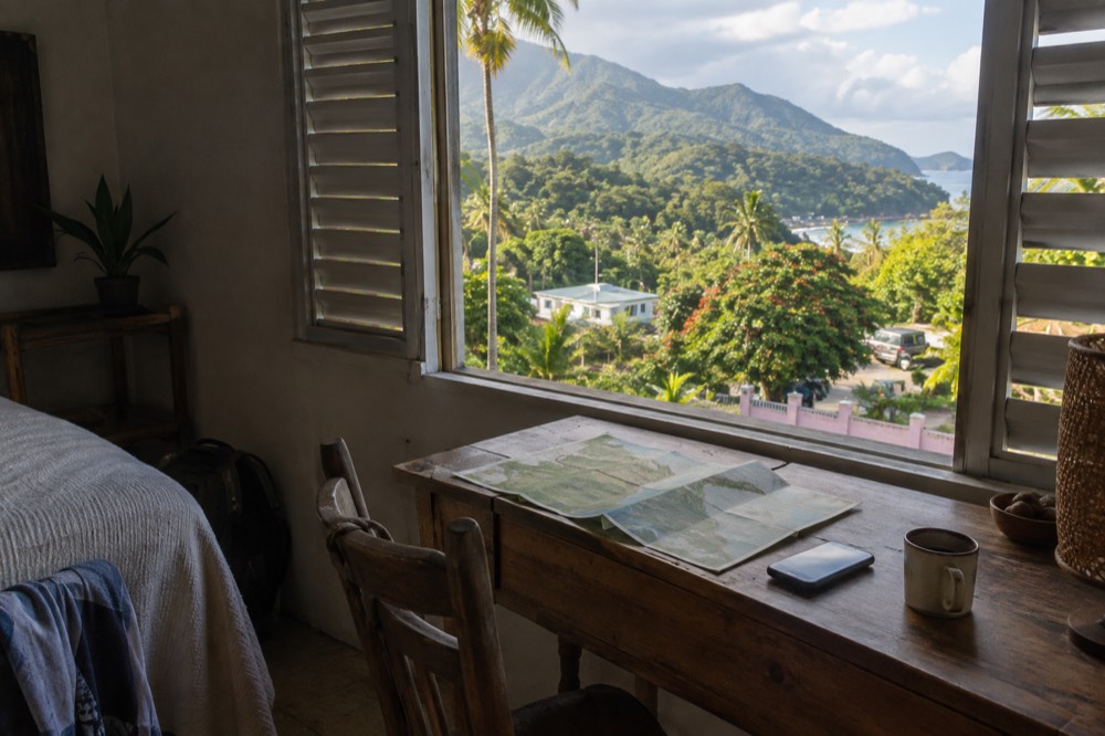 A printed forest map and a smartphone on a hotel desk beside a cup of coffee, with a window showing green hills in the background